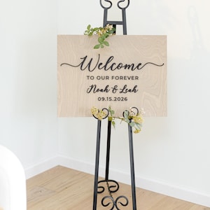 May include: A rectangular light wood sign with visible grain, featuring black text that reads Welcome TO OUR FOREVER Noah & Leah 09.15.2026. The sign is displayed on a black metal easel with scrollwork details and decorated with small green floral sprigs.