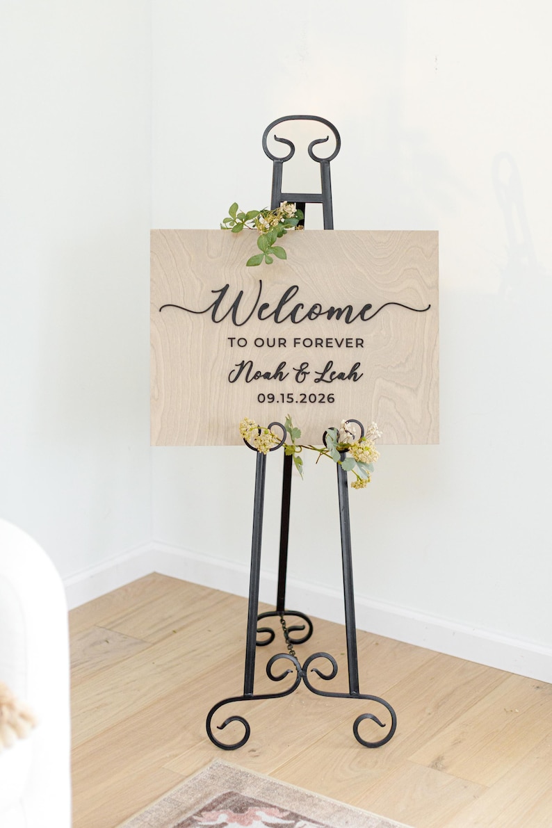 May include: A rectangular light wood sign with visible grain, featuring black text that reads Welcome TO OUR FOREVER Noah & Leah 09.15.2026. The sign is displayed on a black metal easel with scrollwork details and decorated with small green floral sprigs.