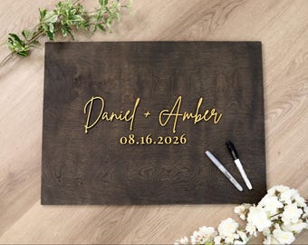 May include: A rectangular dark brown wooden guest book sign featuring the gold-colored raised text Daniel + Amber and the date 08.16.2026. Two markers, one black and one silver, are placed on the surface of the wooden board for signing purposes.
