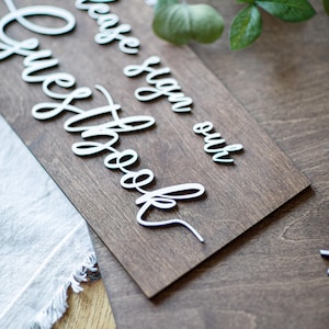 a wooden sign with a cursive message that reads "Please sign our guestbook" placed on a wooden surface.