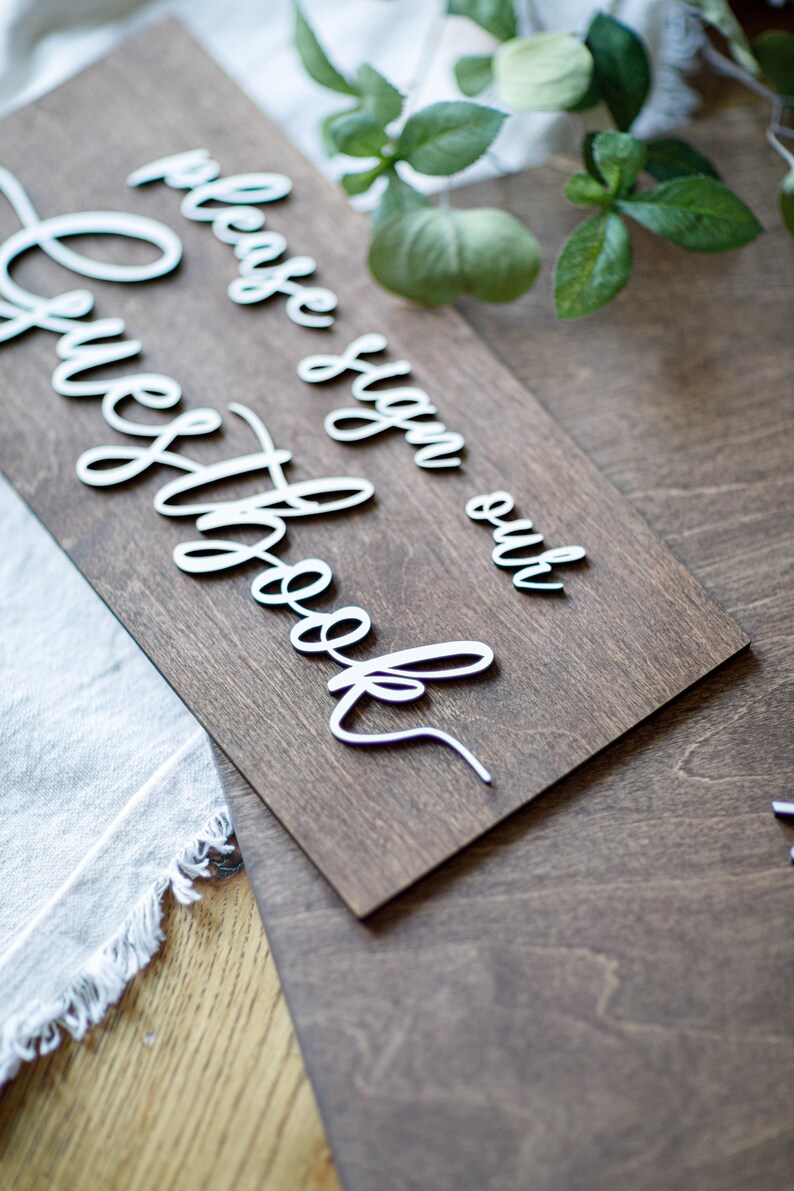 a wooden sign with a cursive message that reads "Please sign our guestbook" placed on a wooden surface.