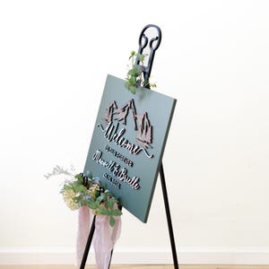 May include: A dark green rectangular wedding welcome sign on a black metal easel. The sign features a 3D wooden mountain graphic, the text "Welcome to our forever", "Maxwell & Brielle", and "05.15.2026". It is decorated with small green floral sprigs.