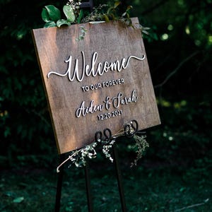 May include: A rectangular wooden sign with a dark brown stained finish, displayed on a black metal easel. The sign features white 3D lettering that reads Welcome TO OUR FOREVER Aiden & Sarah 12.20.2021, accented with small green foliage.