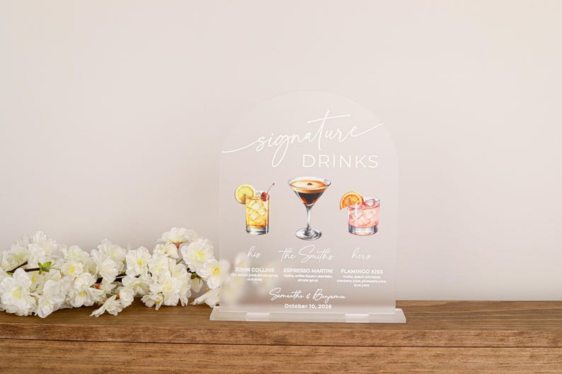 May include: An arched, frosted acrylic sign titled Signature Drinks featuring watercolor illustrations of three cocktails. The text includes John Collins, Espresso Martini, Flamingo Kiss, The Smiths, Samantha & Benjamin, and October 10, 2026.