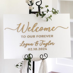 May include: A rectangular white sign with gold lettering reading Welcome TO OUR FOREVER Logan & Taylor 02.10.2024, displayed on a black metal stand decorated with white flowers, green leaves, and white fabric draped at the base.