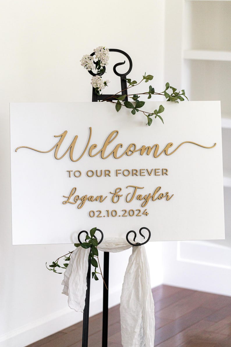 May include: A rectangular white sign with gold lettering reading Welcome TO OUR FOREVER Logan & Taylor 02.10.2024, displayed on a black metal stand decorated with white flowers, green leaves, and white fabric draped at the base.