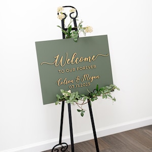 May include: A rectangular olive green sign with gold 3D lettering that reads Welcome TO OUR FOREVER Colson & Megan 07.11.2025. The sign is displayed on a black metal easel decorated with small white flowers and green leaves.