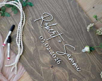 May include: A rectangular dark brown wooden sign featuring white 3D lettering that reads Robert + Sammi and the date 05.02.2026. The sign is accompanied by two black markers, a length of white braided rope, and small green floral accents.