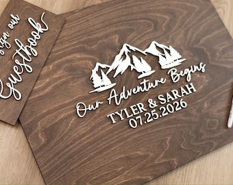 May include: A dark brown wooden guestbook sign with white 3D lettering that reads Please sign our Guestbook and Our Adventure Begins, Tyler & Sarah, 07.25.2026, featuring a mountain and pine tree graphic, accompanied by two markers.