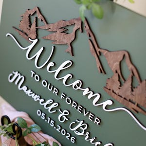 May include: A rectangular olive green sign featuring a dark brown wooden mountain and forest cutout at the top. The white 3D lettering reads "Welcome TO OUR FOREVER Maxwell & Brielle 05.15.2026" in a combination of script and block fonts.
