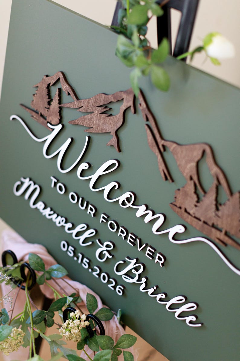 May include: A rectangular olive green sign featuring a dark brown wooden mountain and forest cutout at the top. The white 3D lettering reads "Welcome TO OUR FOREVER Maxwell & Brielle 05.15.2026" in a combination of script and block fonts.