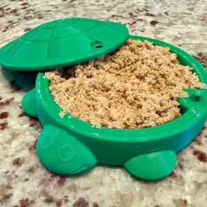 May include: A green turtle-shaped container with a lid, filled with brown sugar. The lid is partially open, revealing the contents. The container has a round body with a turtle shell-shaped lid and turtle feet.