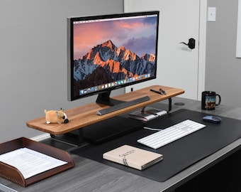 May include: A wooden monitor stand with a black metal frame, a black mouse pad, a white keyboard, a small notebook, and a stuffed animal on a gray desk.