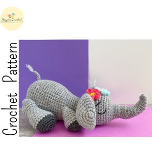 Puede incluir: Un juguete de elefante de crochet gris con flores rosas y azules en la cabeza, acostado sobre una superficie morada y rosa. El elefante tiene un ojo cerrado y una pequeña sonrisa.