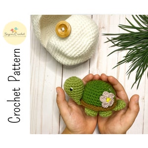 Może przedstawiać: Zielona szydełkowa żółw z brązowym pancerzem i różowym kwiatem na grzbiecie. Żółw jest trzymany w dłoniach osoby.