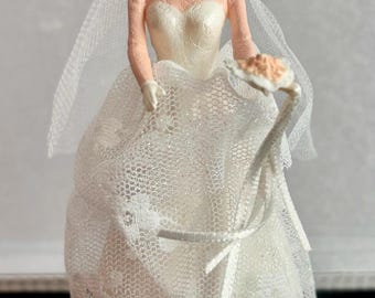 Puede incluir: Una figura de novia de porcelana blanca con un vestido de encaje blanco y velo, sosteniendo un ramo de flores.