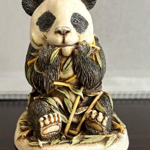 May include: A small ceramic figurine of a panda bear sitting and eating bamboo. The panda is black and white with a brown nose and black eyes. The figurine is surrounded by green leaves and yellow bamboo stalks. The base of the figurine is cream colored and has the number 1000 on it.