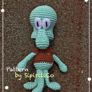 Könnte beinhalten: Ein grüner gehäkelter Oktopus mit braunem Hemd und weißen Augen. Der Oktopus liegt auf einer braunen Oberfläche. Der Text "Pattern by Sipirilico" ist unter dem Oktopus sichtbar.