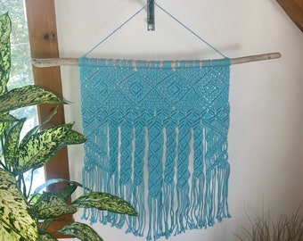 May include: A light blue macrame wall hanging with a wood branch at the top and fringe at the bottom.