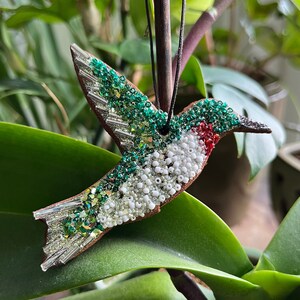 May include: A green, white, and red beaded hummingbird ornament hanging from a branch. The hummingbird is made of beads and has a long, thin beak.