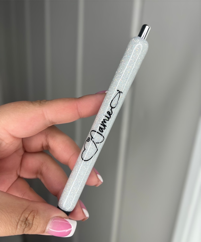 May include: A white glitter pen with a silver top and black ink. The pen has a stethoscope symbol and the name "Jamie" printed on it.