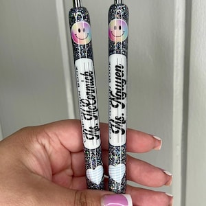 May include: Two black glitter pens with a smiley face design. The pens have a white label with the text "Composition Book" and the teacher's name. One pen says "Mrs. Goldsmith" and the other says "Mr. Haugen".