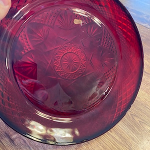 May include: A ruby red glass plate with a diamond pattern around the rim and a starburst design in the center. The plate has a slightly raised edge and is made of a transparent material.
