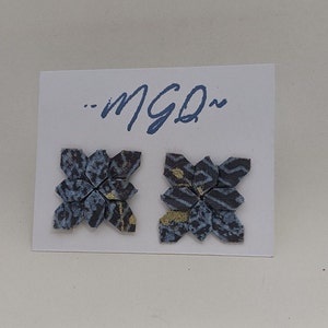 May include: A pair of handmade stud earrings with a floral design. The earrings are made of folded paper in shades of blue, black, and gold, and are mounted on a white card. The card has the handwritten text "...MGD~" in blue.