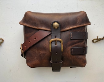 May include: A brown leather satchel bag with a brass buckle and a strap. The bag has a flap with a brass button closure.