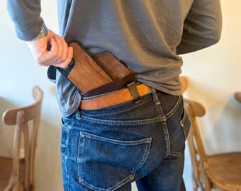 May include: A dark brown leather holster case containing a smartphone with a wood-grain back panel. The holster is secured to a tan leather belt worn over dark blue denim jeans. The device is partially visible, showing a smooth, polished finish.