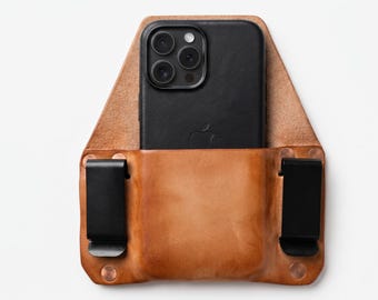 May include: A tan leather holster featuring a central pocket holding a black smartphone with a triple-lens camera system. The holster includes two black metal belt clips attached with copper-coloured rivets on the left and right sides.