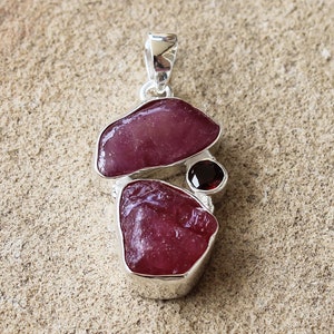 May include: A silver pendant featuring three pink and red gemstones, including a round garnet, set in a silver frame.