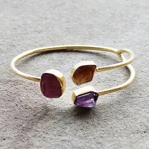 Pode incluir: Pulseira de ouro com três pedras preciosas de cores diferentes: uma rosa, uma roxa e uma marrom.