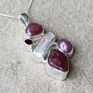 May include: A silver pendant necklace with a cluster of gemstones. The pendant features deep red rubies, a rectangular pearl, and a small red garnet. The gemstones are set in a silver frame.
