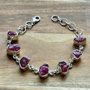 Könnte beinhalten: Ein silbernes Armband mit rohen, tiefroten Rubinsteinen. Die Steine sind in Silberfassungen gefasst und durch silberne Kettenglieder verbunden. Das Armband hat einen Karabinerverschluss. Die Edelsteine haben eine natürliche, raue Textur.