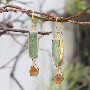 May include: A pair of drop earrings featuring a rectangular, light green stone suspended above a smaller, irregular amber-coloured stone. Both stones are set in gold-toned metal frames and attached to gold-toned ear wires for a dangling effect.