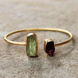 May include: A gold bangle bracelet featuring a green and red gemstone accent. The gemstones are raw and have a natural, irregular shape.