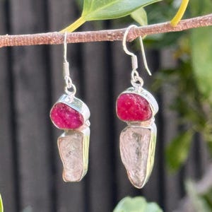 May include: Dangle earrings with a red gemstone above a clear, irregular-shaped stone. The stones are set in silver metal and hang from silver ear wires. The earrings are shown against a blurred natural backdrop. The earrings are a unique piece of jewellery.