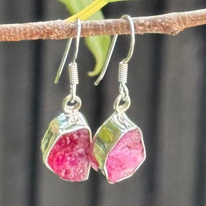 May include: A pair of silver drop earrings with raw, pink-red gemstones. The earrings hang from a simple hook and a coiled spring detail. The gemstones are set in a silver bezel. The background is blurred.