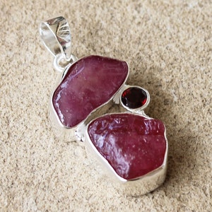 May include: A silver pendant featuring three raw gemstones: two pink rubies and one red garnet. The rubies are irregularly shaped and have a smooth, polished surface. The garnet is round and has a slightly rough texture.