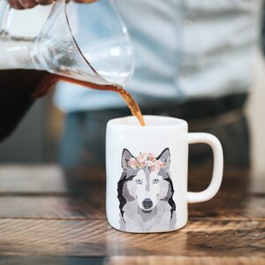 May include: A white ceramic mug with a gray and white wolf wearing a floral crown. Coffee is being poured into the mug from a glass carafe.