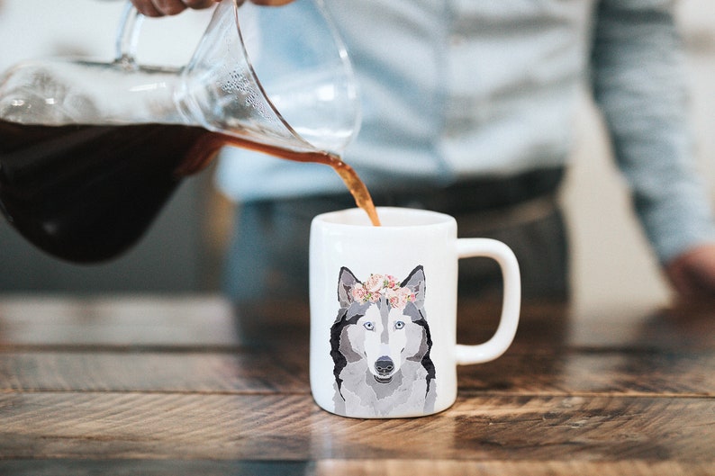 May include: A white ceramic mug with a gray and white wolf wearing a floral crown. Coffee is being poured into the mug from a glass carafe.