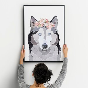 May include: A framed print of a wolf with a floral crown. The wolf is grey and white with blue eyes. The print is in a black frame and is being hung on a white wall.