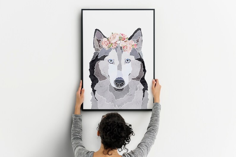 May include: A framed print of a wolf with a floral crown. The wolf is grey and white with blue eyes. The print is in a black frame and is being hung on a white wall.