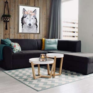 May include: A gray and white wolf with a floral crown is featured in a framed print hanging on a wood wall in a living room. The room has a gray sectional sofa, two white coffee tables, and a teal and white patterned rug.