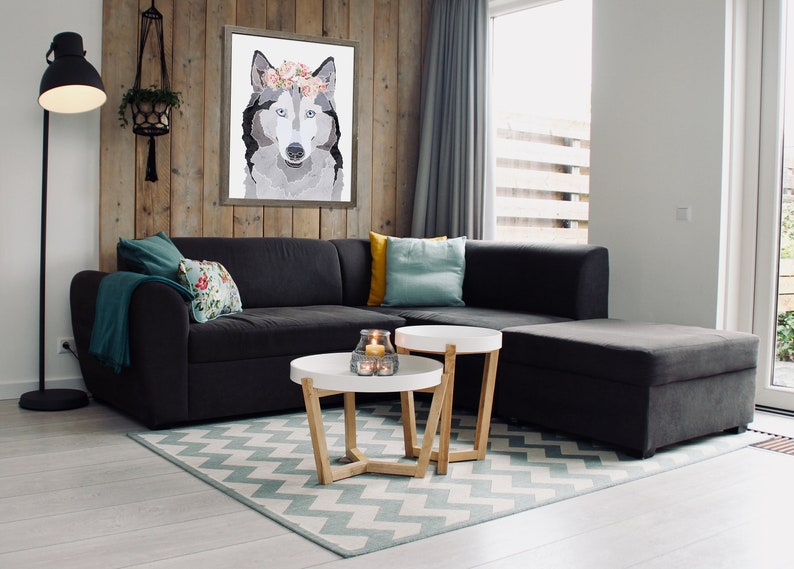 May include: A gray and white wolf with a floral crown is featured in a framed print hanging on a wood wall in a living room. The room has a gray sectional sofa, two white coffee tables, and a teal and white patterned rug.