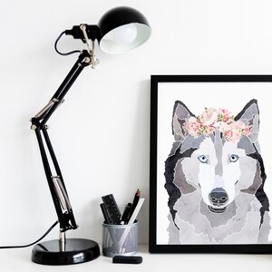 May include: A black and white watercolor illustration of a wolf wearing a pink flower crown. The wolf is looking directly at the viewer. The illustration is framed in a black frame and is hanging on a white wall.