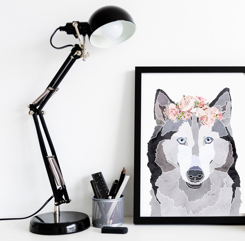May include: A black and white watercolor illustration of a wolf wearing a pink flower crown. The wolf is looking directly at the viewer. The illustration is framed in a black frame and is hanging on a white wall.