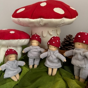 Könnte beinhalten: Eine skurrile Szene mit vier kleinen Puppen in grauen Outfits und roten Pilzhüten mit weißen Punkten. Große rote und weiße Pilzdekorationen befinden sich im Hintergrund und schaffen ein Märchenmotiv. Die Puppen sind auf einem grünen Stoff angeordnet.