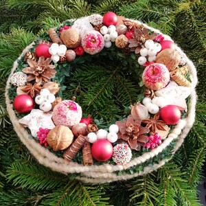 Könnte beinhalten: Ein festlicher Weihnachtskranz mit rustikalem Design. Er hat eine Basis aus grünen Zweigen, verziert mit roten Kugeln, gezuckerten Äpfeln, Tannenzapfen, Walnüssen und weißen Beeren. Der Kranz ist mit einem dicken, beigen Seil umwickelt.